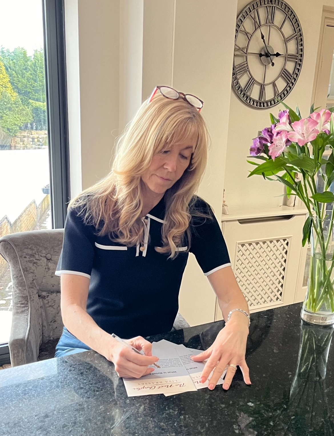A woman with blonde hair sits at a table writing on branded stationery, with flowers in a vase beside her and a large wall clock in the background.