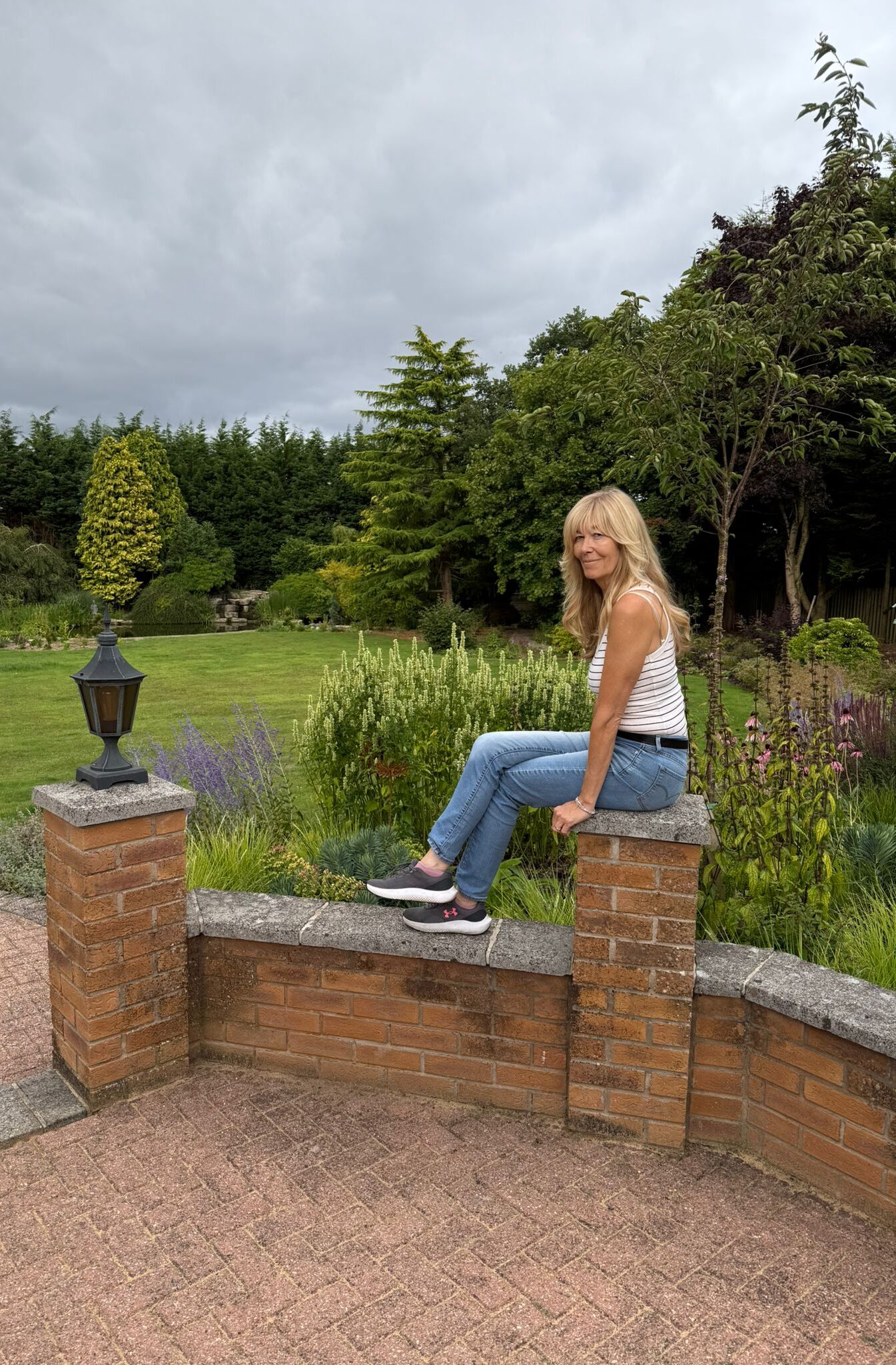 A woman with long blonde hair, dressed in a white striped top and blue jeans, sits on a brick garden wall surrounded by lush greenery and blooming flowers under a cloudy sky.